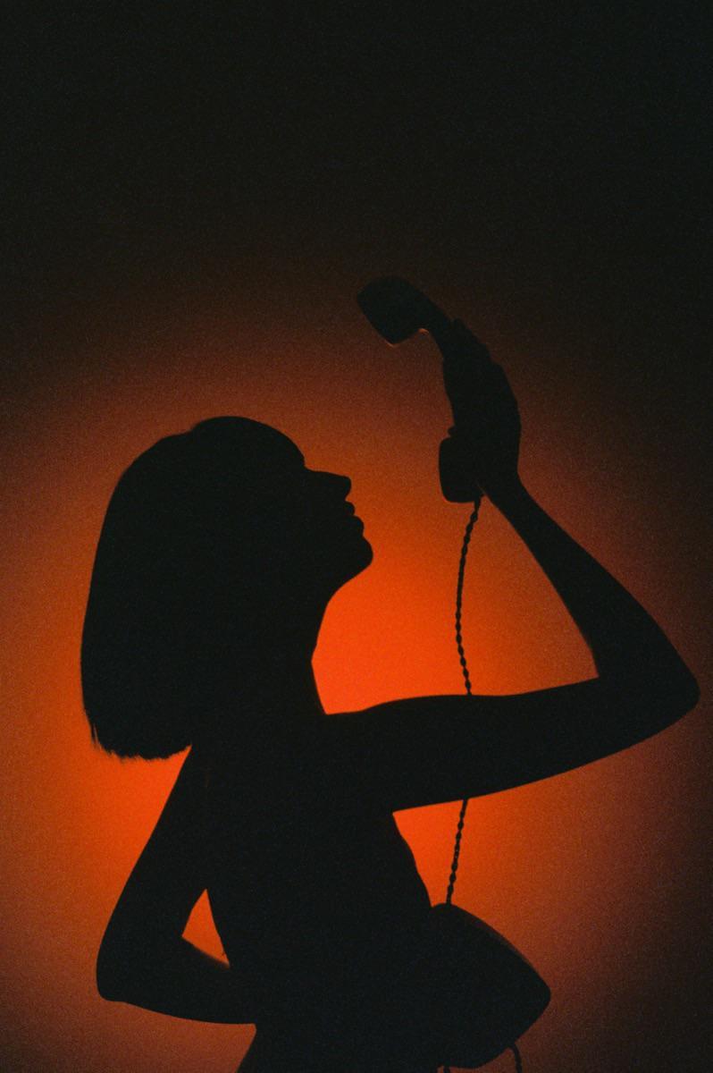 A woman in silhouette holds a telephone receiver to her ear against an orange background.