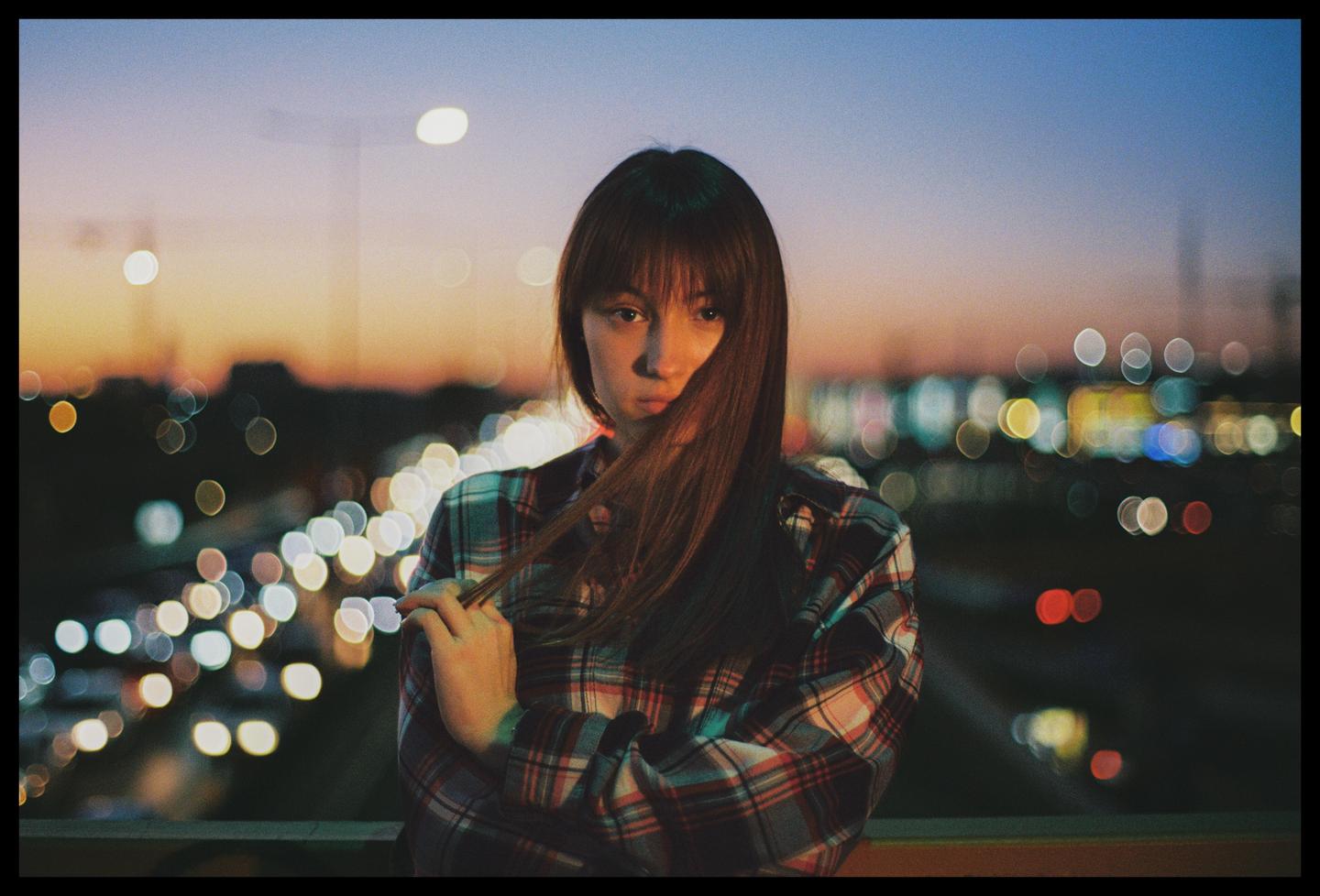 A young woman with long hair and a plaid shirt stands in front of a blurred city lights at dusk.