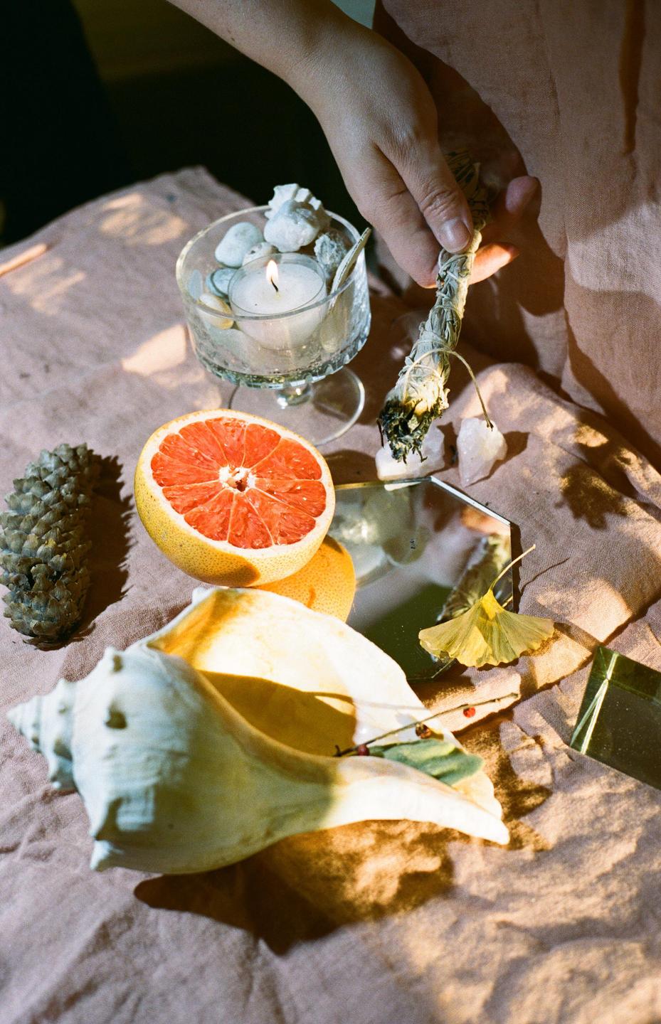 A hand holds a burning sage smudge stick over a collection of objects including a grapefruit, seashell, pinecone, and candle.