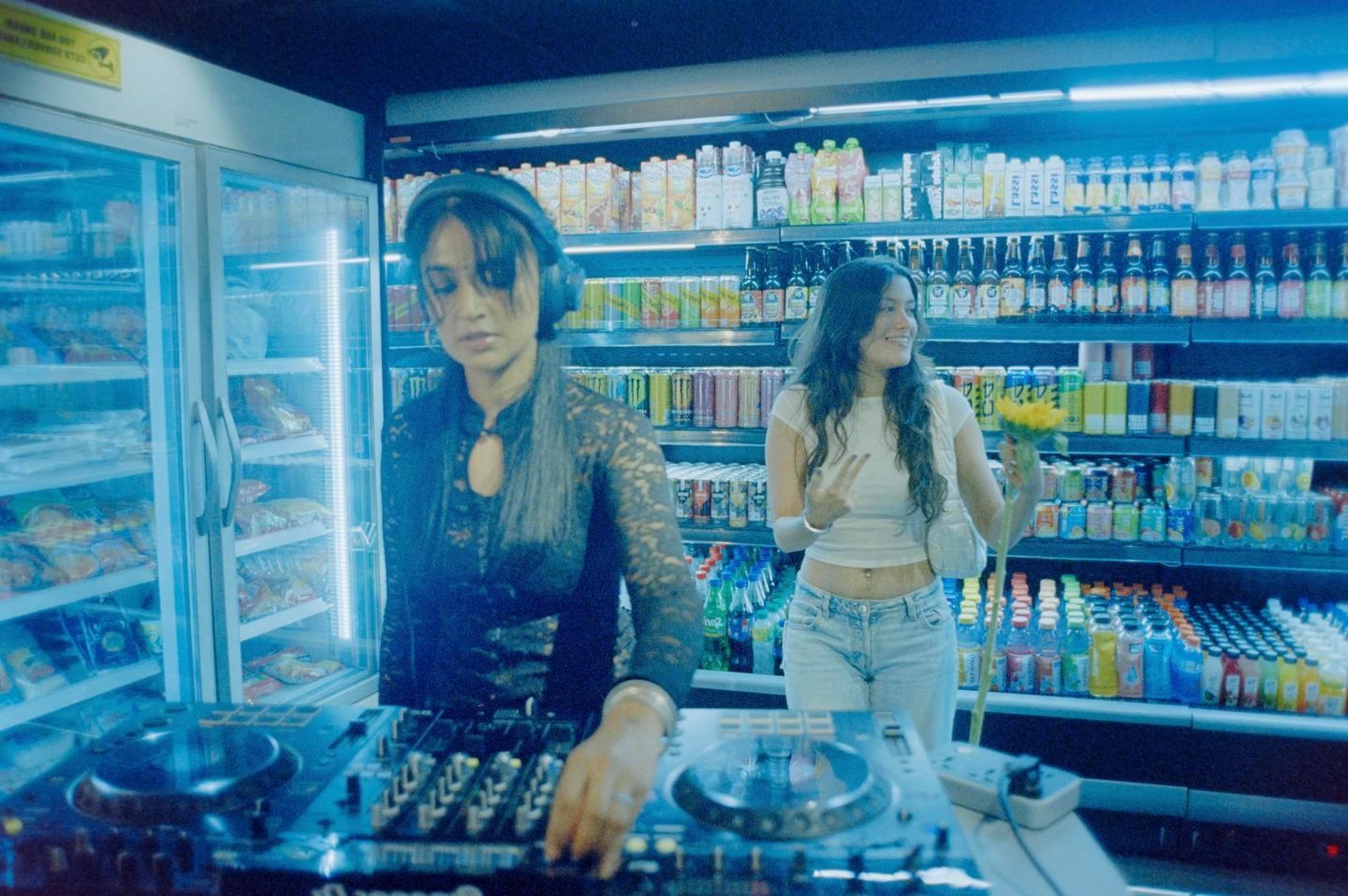 A woman wearing headphones is DJing in a convenience store while another woman holds a sunflower and makes a peace sign.