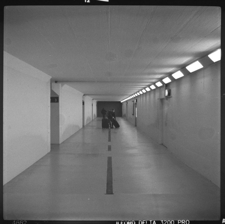 Two people with luggage stand in a long, brightly lit tunnel.