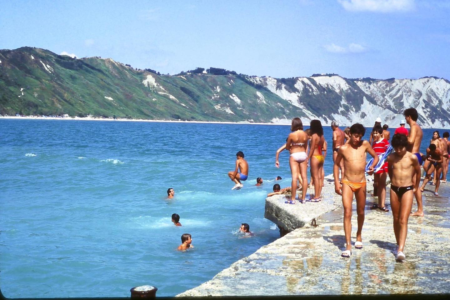 A group of people are swimming and standing on a concrete pier next to a body of water with mountains in the background.