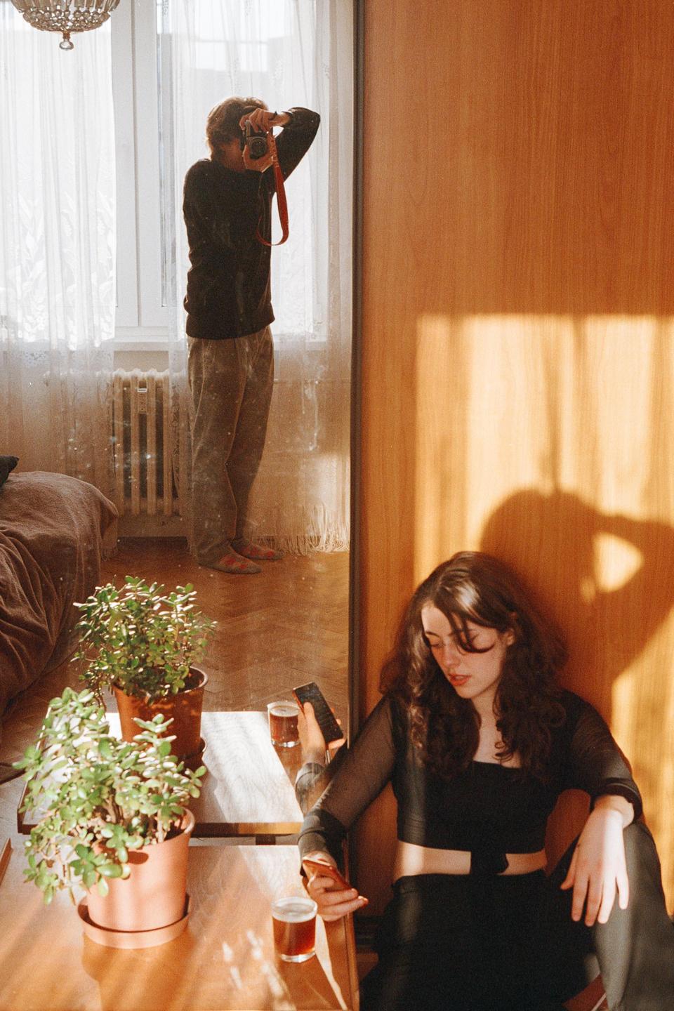 A man takes a self-portrait in a mirror while a woman sits next to a table with two potted plants and two glasses.