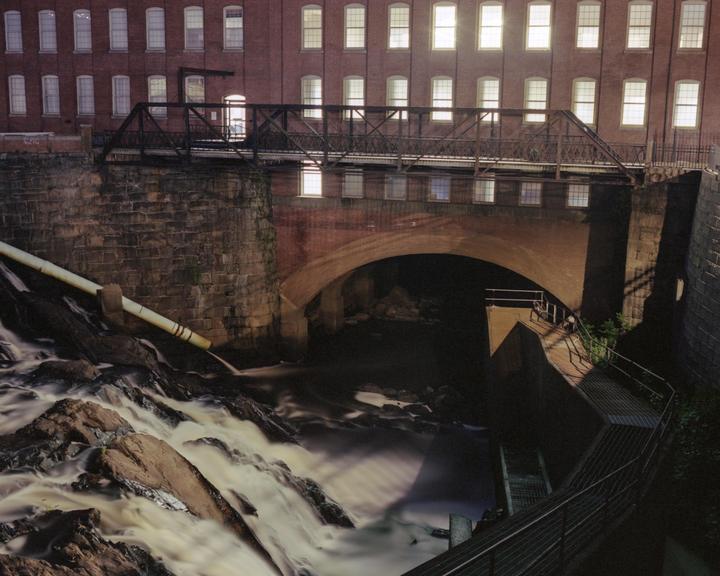 A waterfall flows under a bridge and a brick building with many illuminated windows at night.