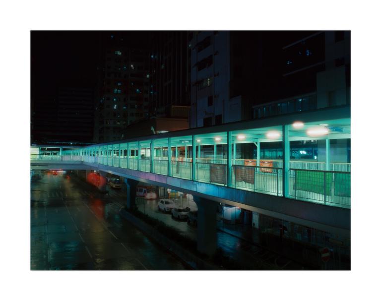 A long, illuminated pedestrian bridge stretches over a wet street with cars and buildings in the background at night.