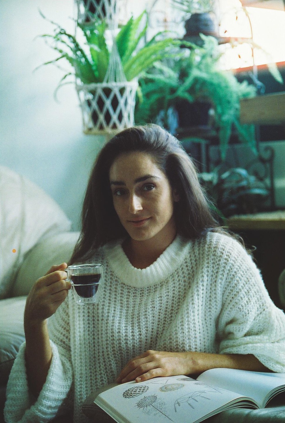 A woman in a white sweater holds a cup of coffee while looking at a book.