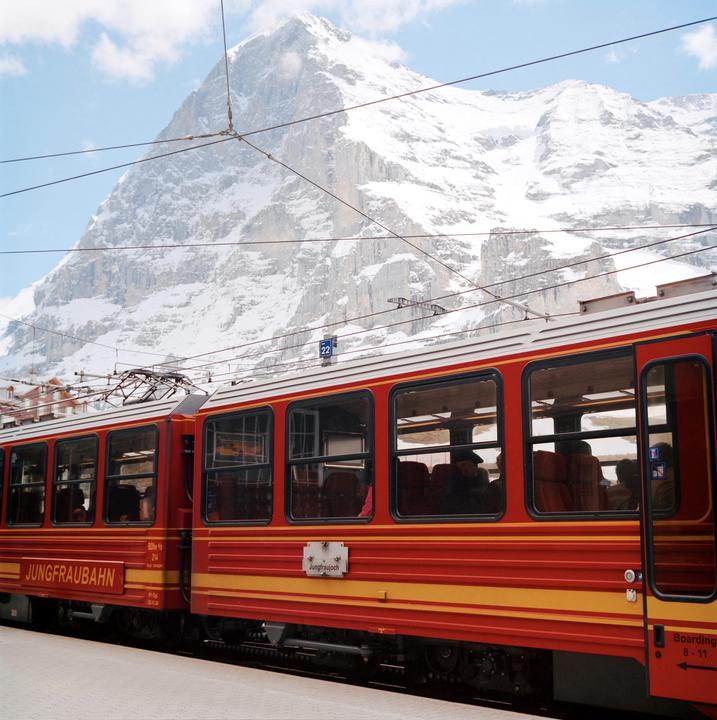 A red and yellow train is parked at a station with a snow-covered mountain in the background.