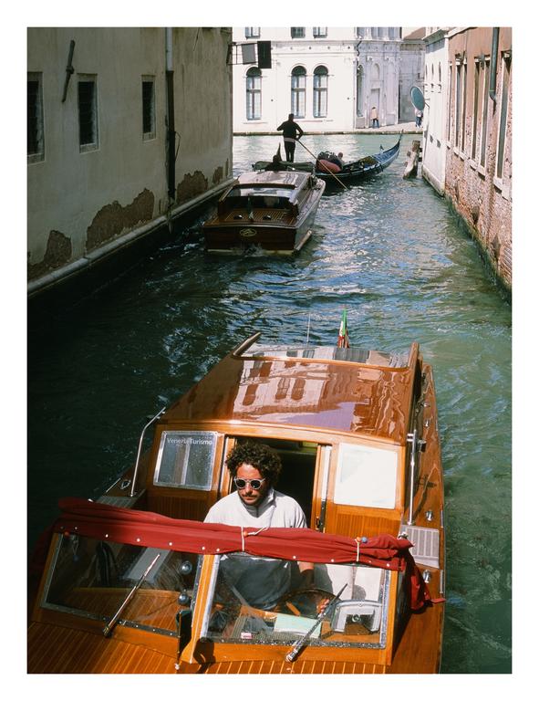A man in sunglasses drives a water taxi down a canal in Venice, with a gondola and another boat visible behind him.