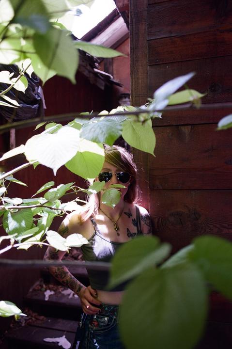 A woman with sunglasses and tattoos stands partially obscured by green leaves next to a wooden wall.