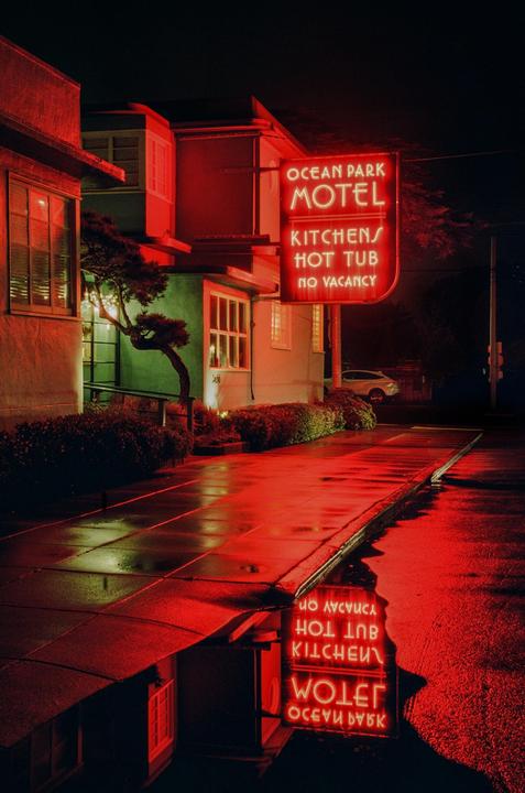 A neon motel sign reflects in a puddle on a wet sidewalk at night.