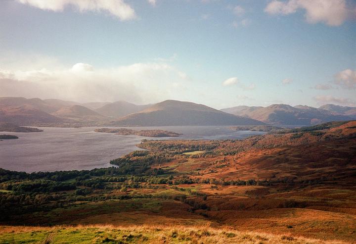 A wide view shows a lake surrounded by mountains and autumn foliage under a hazy sky.