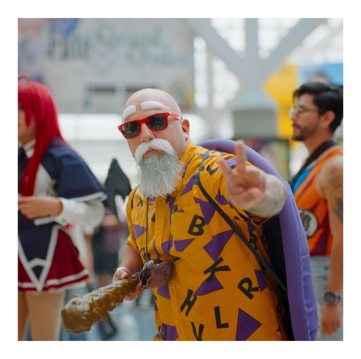 A man in a yellow shirt, red sunglasses, and a white beard makes a peace sign while holding a staff.