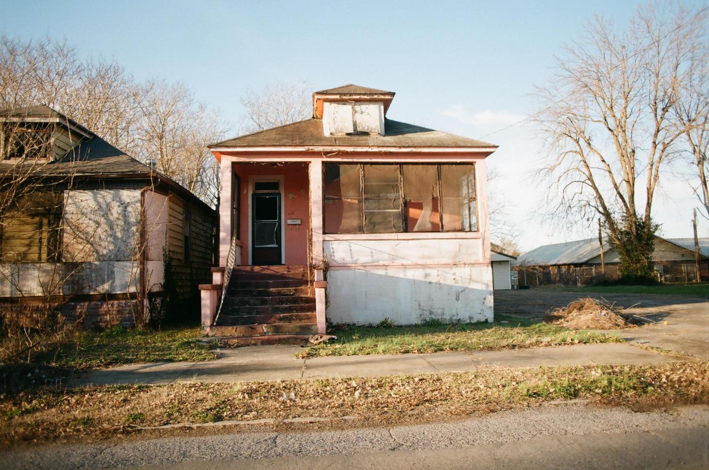 A dilapidated pink house with a screened-in porch stands next to another rundown building under a clear sky.