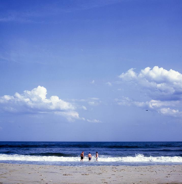 Three men stand in the shallow water of a beach with waves breaking around them under a blue sky with clouds.