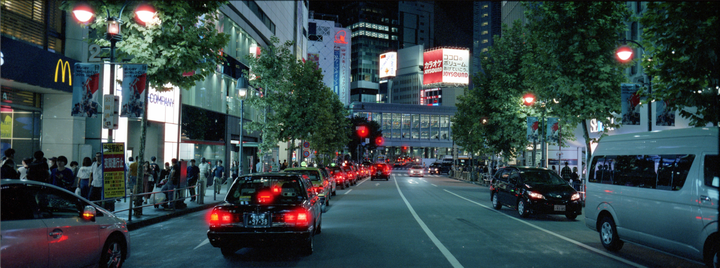 A street scene at night shows a line of cars with red taillights, people on the sidewalk, and illuminated buildings with signs.