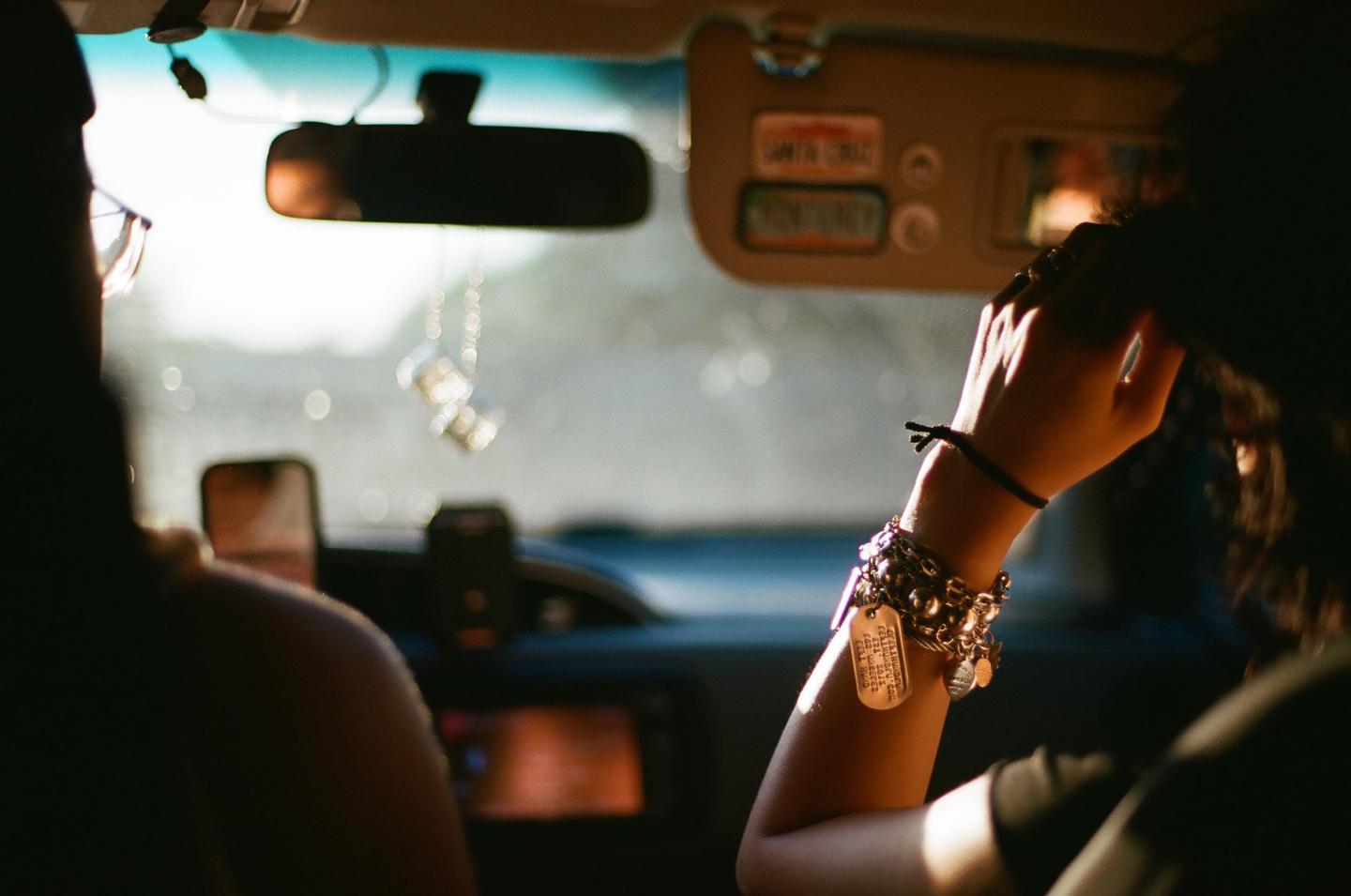 A person with a bracelet on their arm is sitting in the passenger seat of a car, with a pair of fuzzy dice hanging from the rearview mirror.