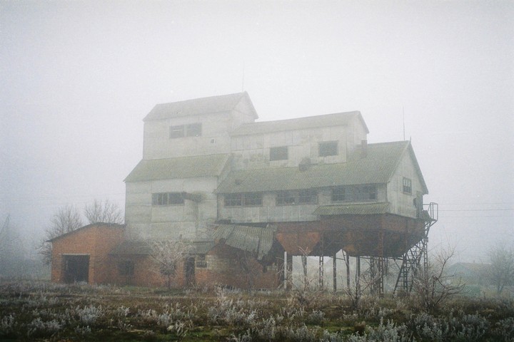 An old industrial building with a green roof stands in a foggy, desolate landscape with frosted vegetation in the foreground.
