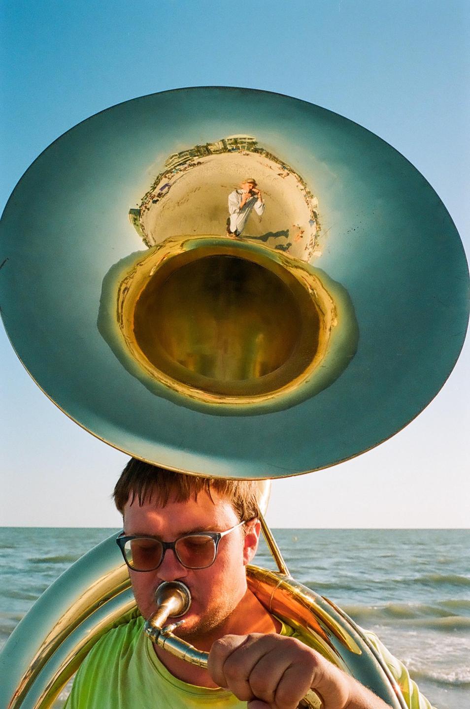 A man plays a tuba on the beach, with a reflection of a person taking a photo visible in the instrument's bell.