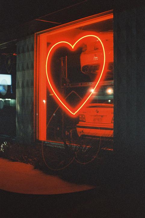 A neon heart sign glows red in a window, reflecting on a car and bicycle below.