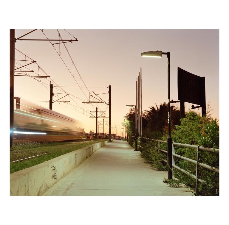 A blurred train speeds past a concrete walkway lined with streetlights and green bushes under a pale orange sky.