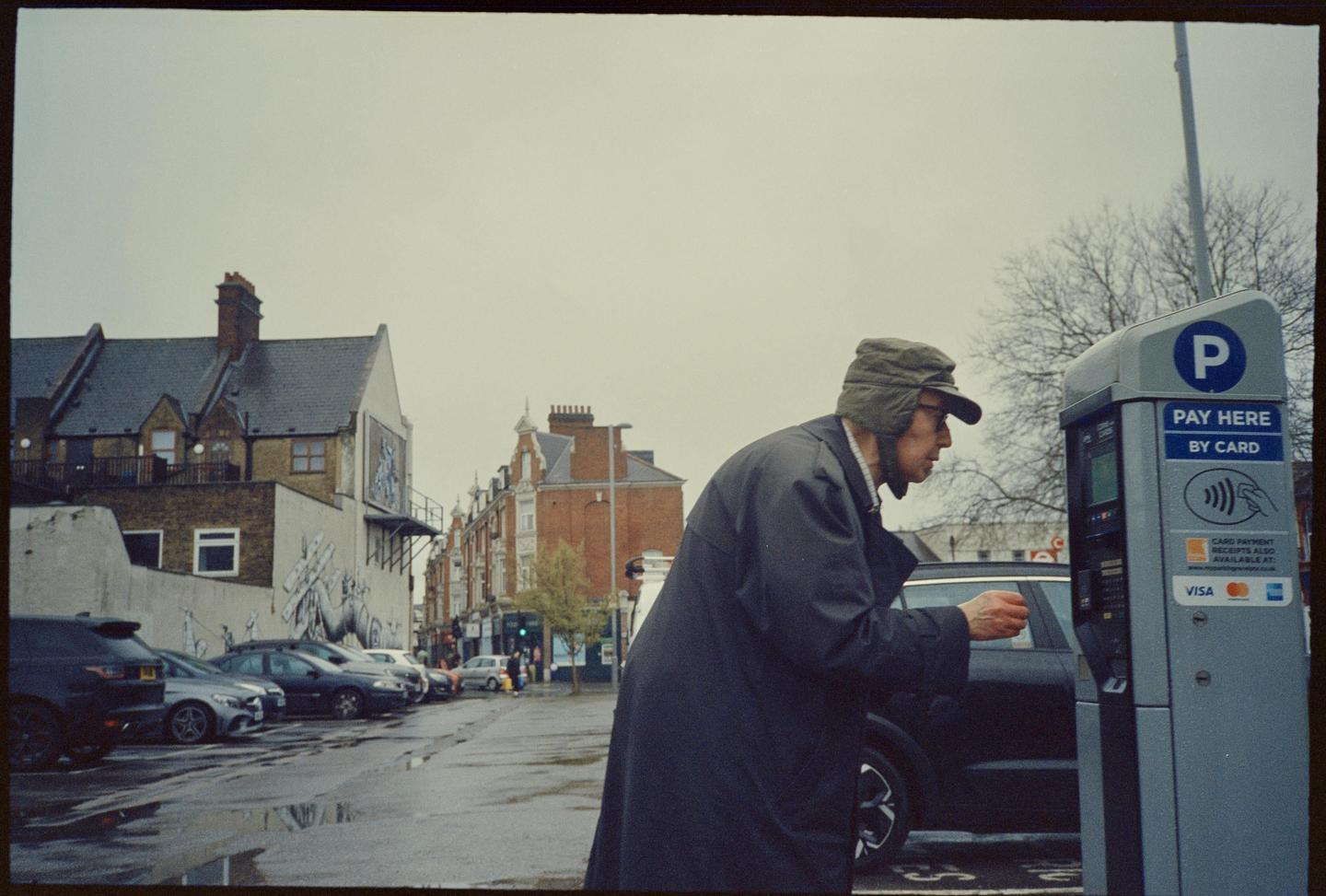 A man in a coat and hat stands next to a parking meter on a street with parked cars and buildings.