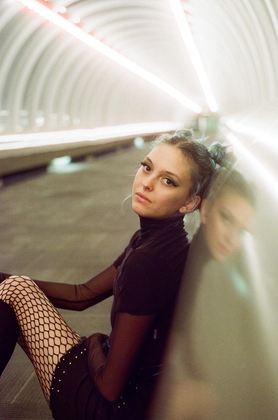 A woman with blue hair buns and fishnet stockings sits on the ground in a tunnel with bright lights.