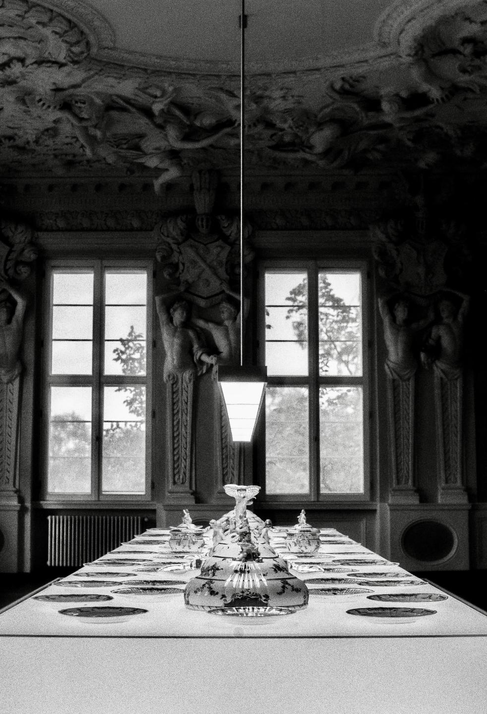 A long dining table set with plates and glassware sits in a grand room with ornate ceilings and large windows.
