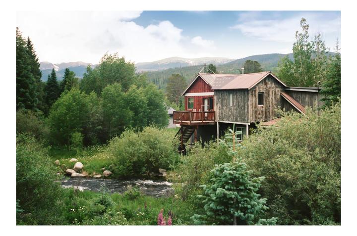 A rustic house with a red roof stands among lush green trees and a flowing river, with mountains in the background.