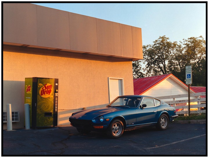 A blue car is parked next to a Sun Drop vending machine outside a building with a red roof.