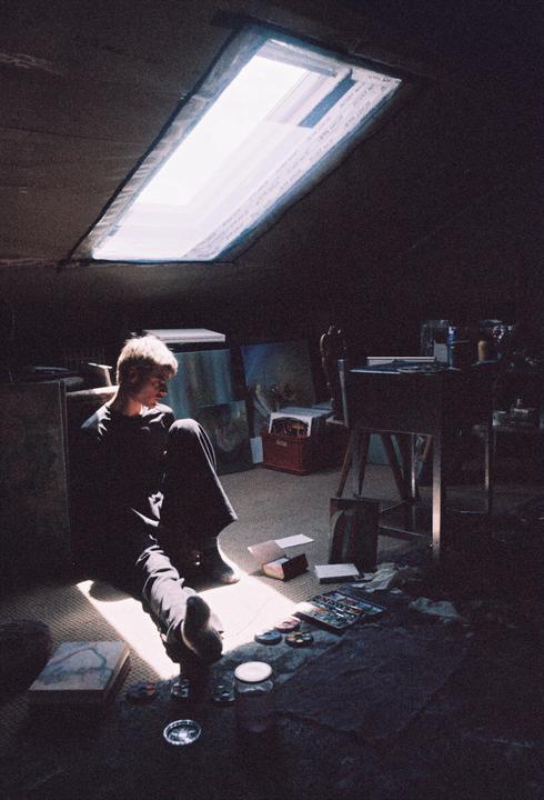 A man sits on the floor in a dimly lit attic studio, illuminated by a skylight.