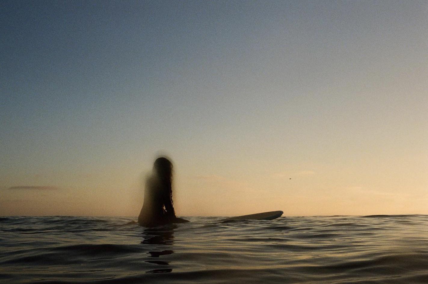 A surfer sits on a surfboard in the ocean at sunset.