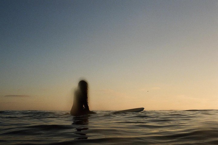 A surfer sits on a surfboard in the ocean at sunset.
