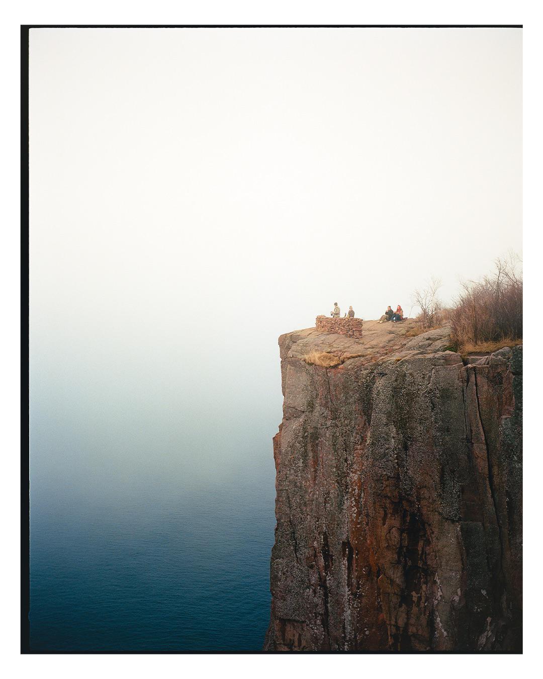 A group of people sit on a rocky cliff overlooking a body of water under a hazy sky.