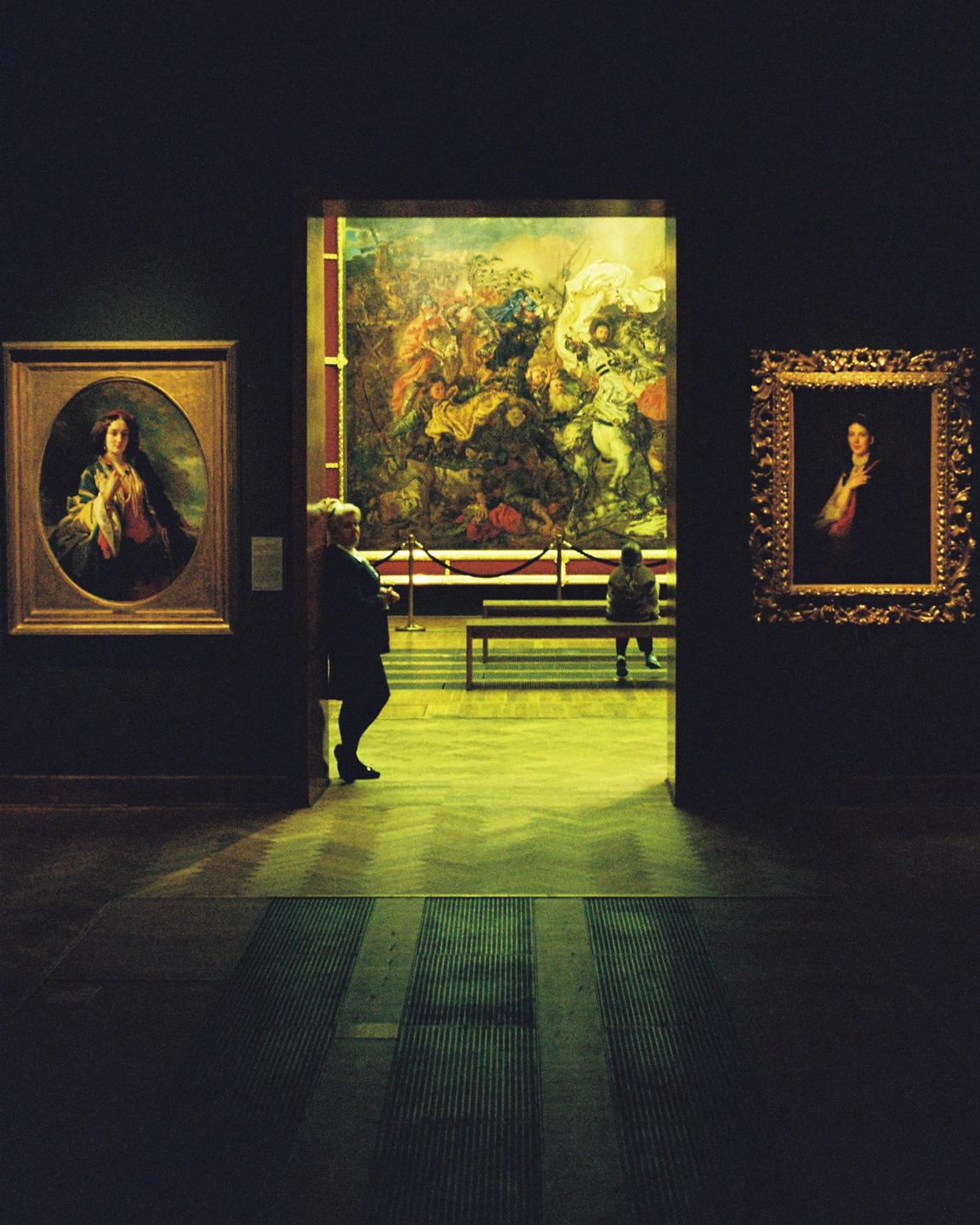 A person stands in a dark museum gallery looking at a large painting, while another person sits on a bench in the background.