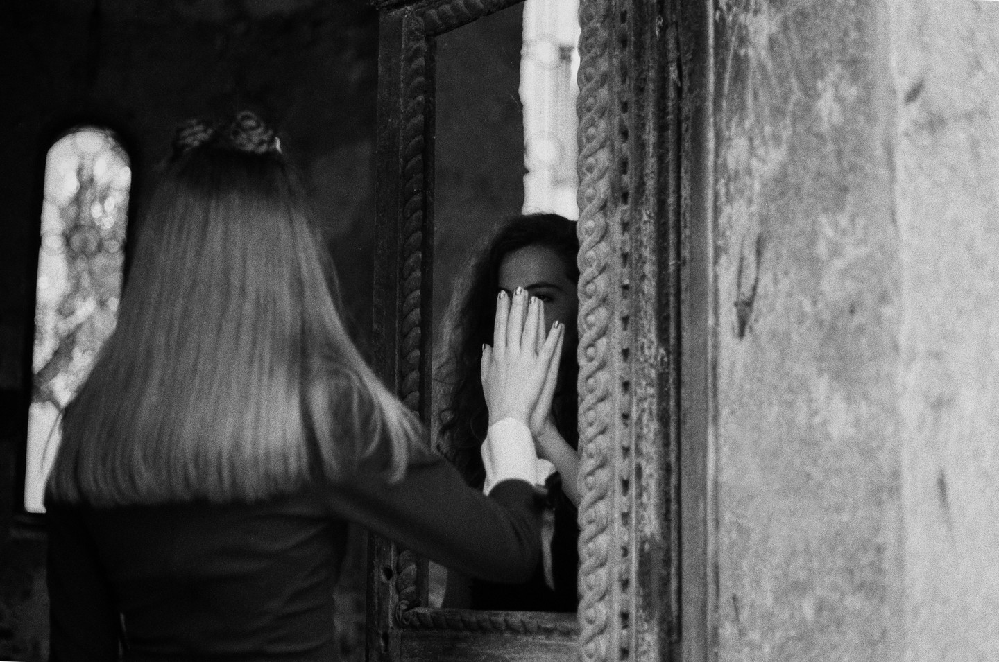 A woman with long hair stands with her back to the viewer, looking at her reflection in a mirror where her hands cover her face.