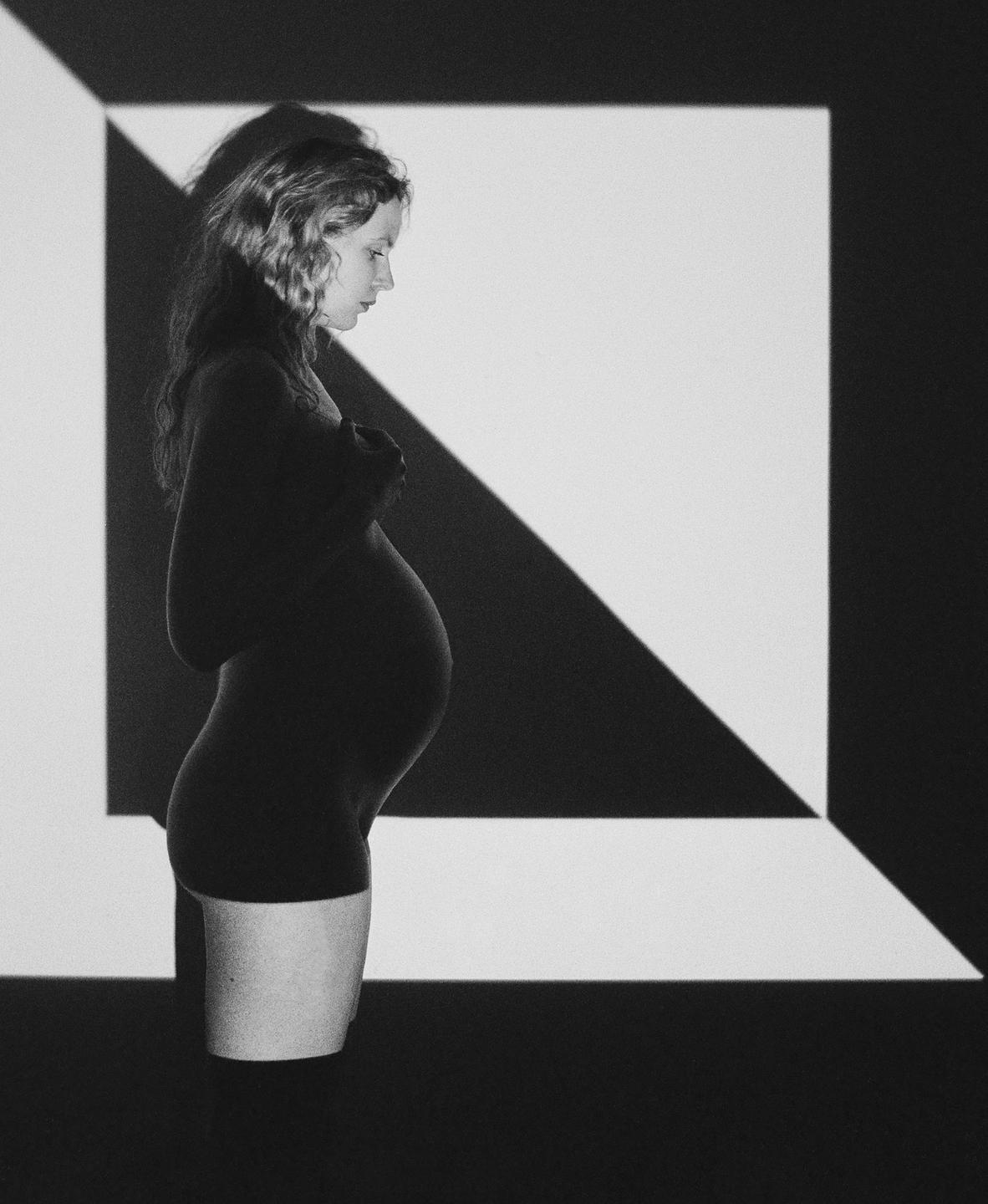 A pregnant woman stands in profile against a wall with geometric light patterns.