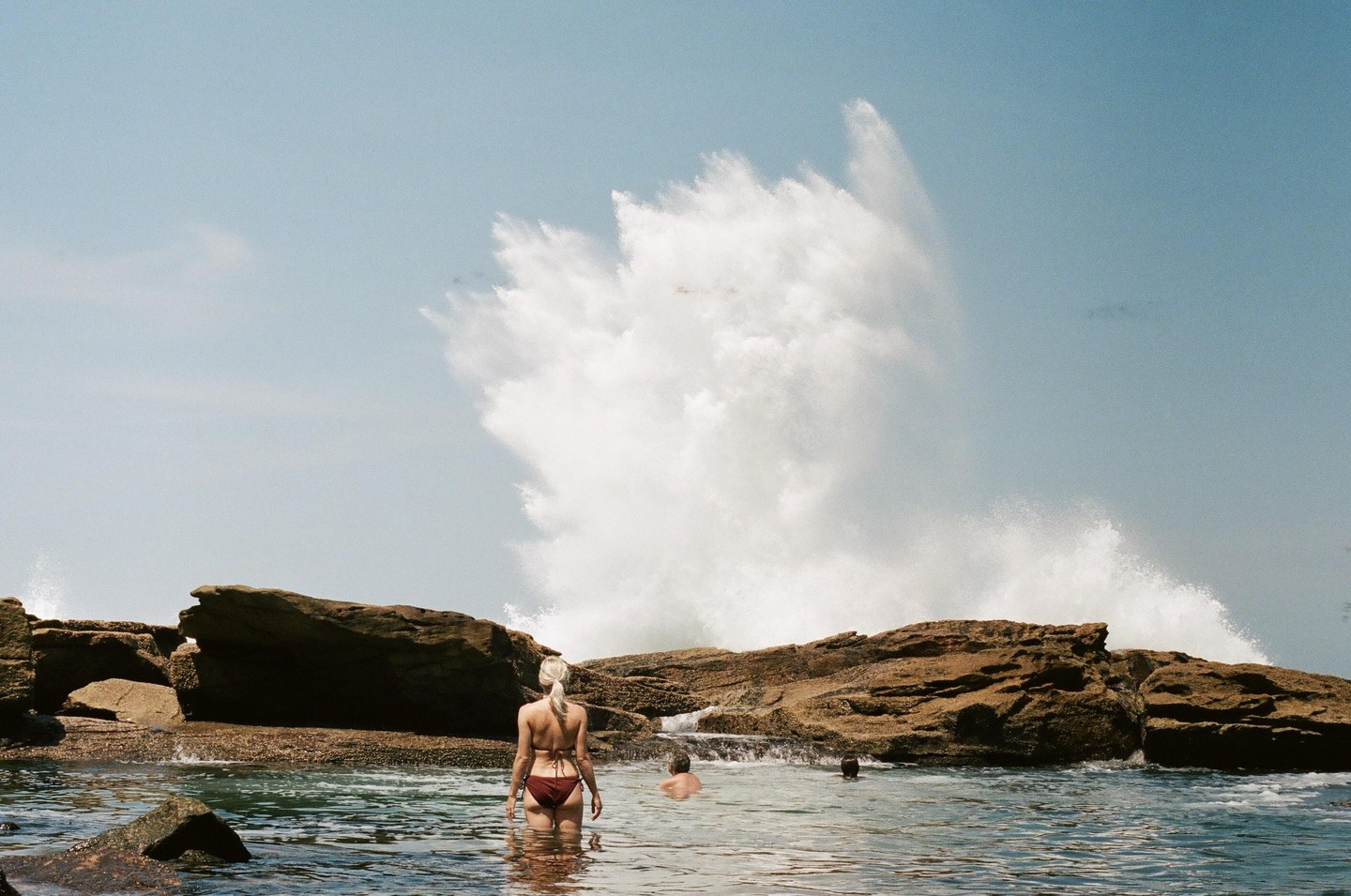 A woman in a bikini stands in the ocean with two other people swimming near large rocks as a huge wave crashes in the background.
