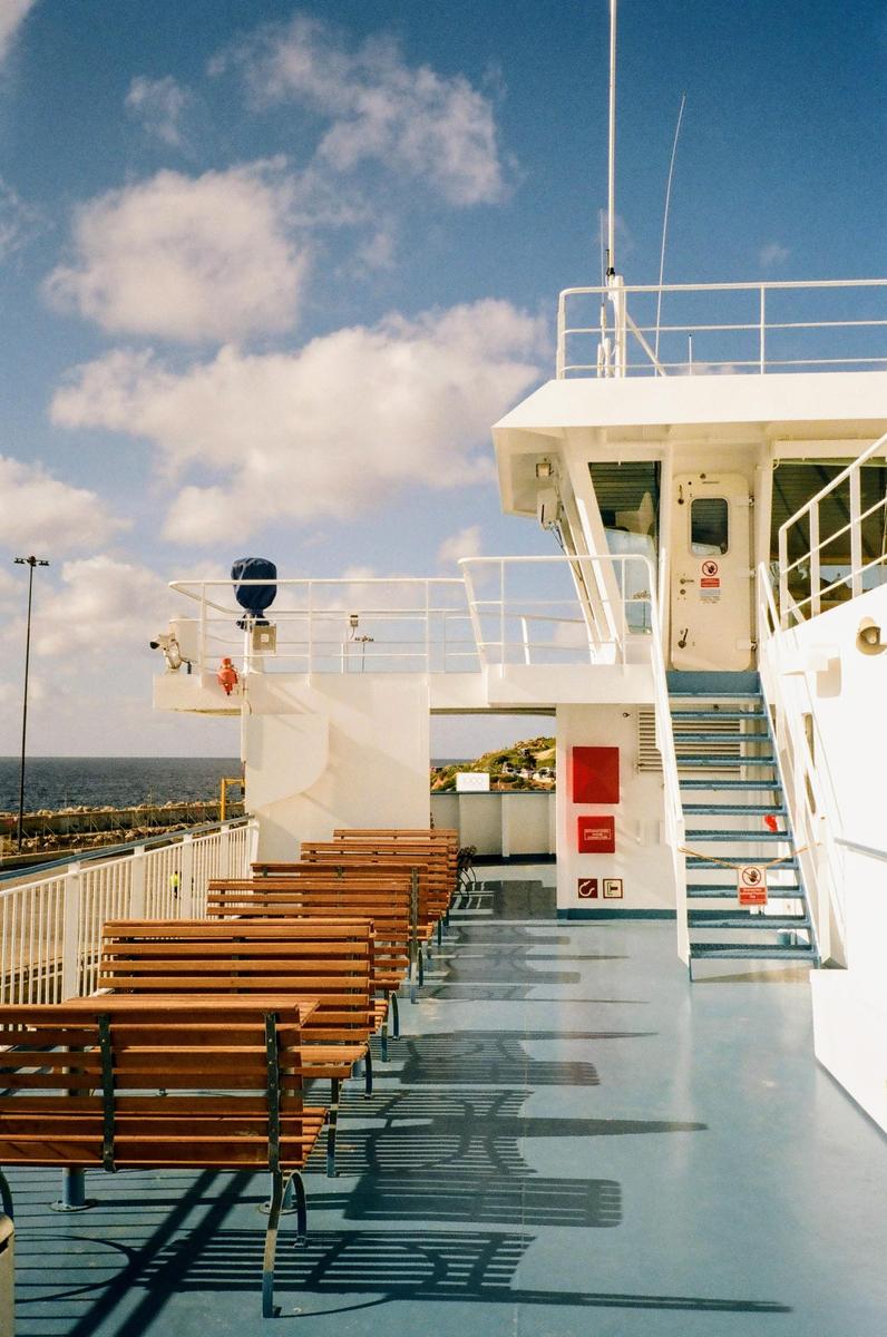 A row of wooden benches casts long shadows on the deck of a ferry under a blue sky with clouds.