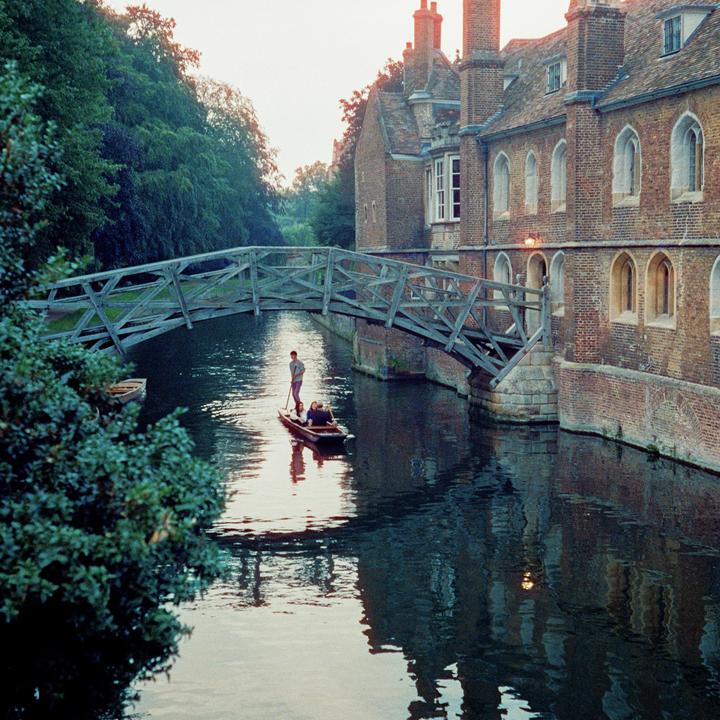 A man punts a boat with two people under a wooden bridge next to a brick building.