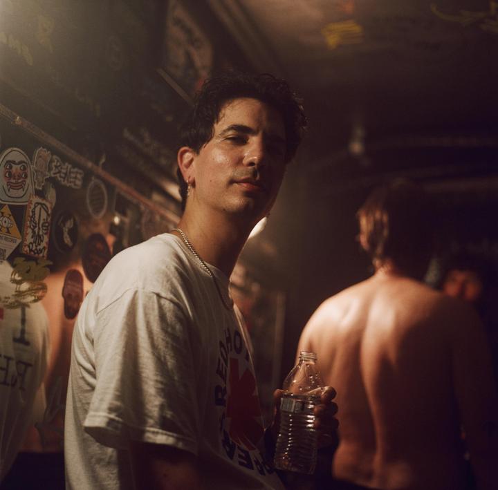 A man in a white t-shirt holds a water bottle and looks at the camera while another man stands with his back to the camera in a dimly lit room.