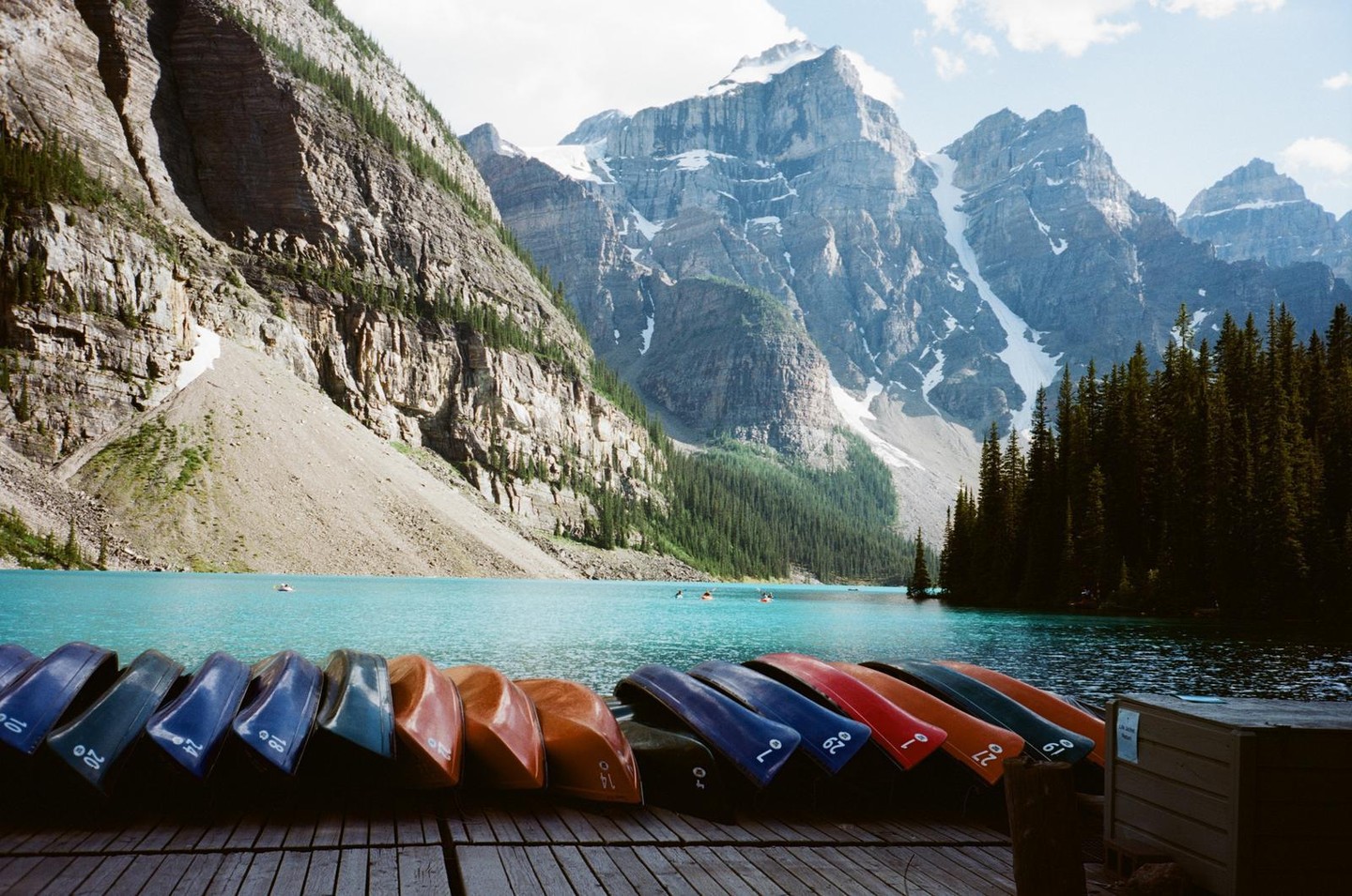 Colorful canoes are lined up on a wooden dock in front of a turquoise lake surrounded by mountains and a pine forest.