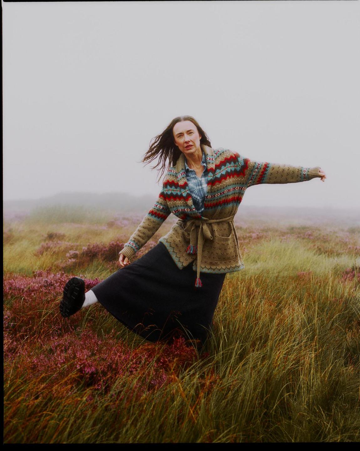 A woman in a patterned cardigan and long skirt stands in a field of tall grass and purple flowers, with a hazy sky in the background.
