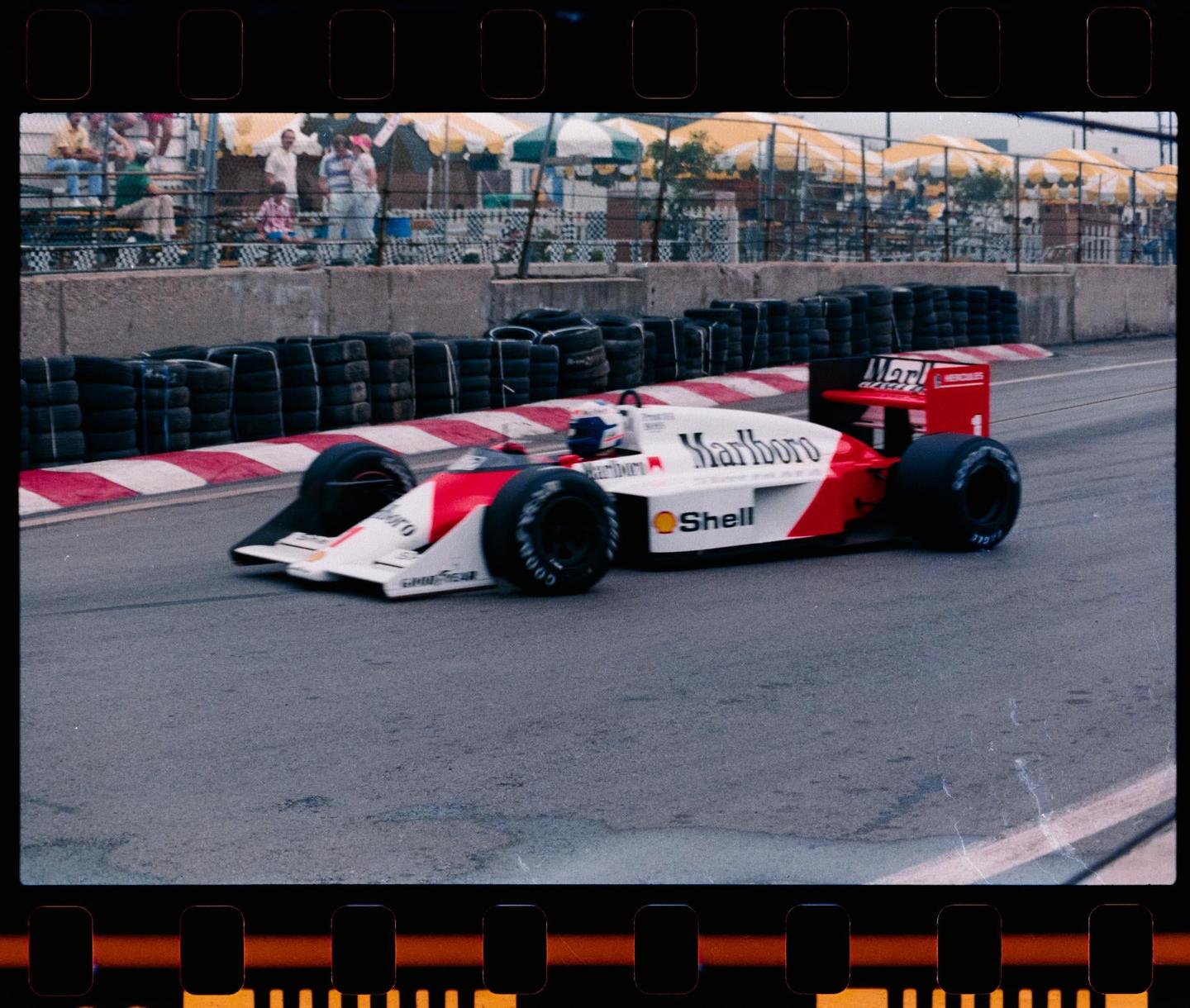 A red and white Formula 1 race car is speeding on a track with spectators and umbrellas in the background.
