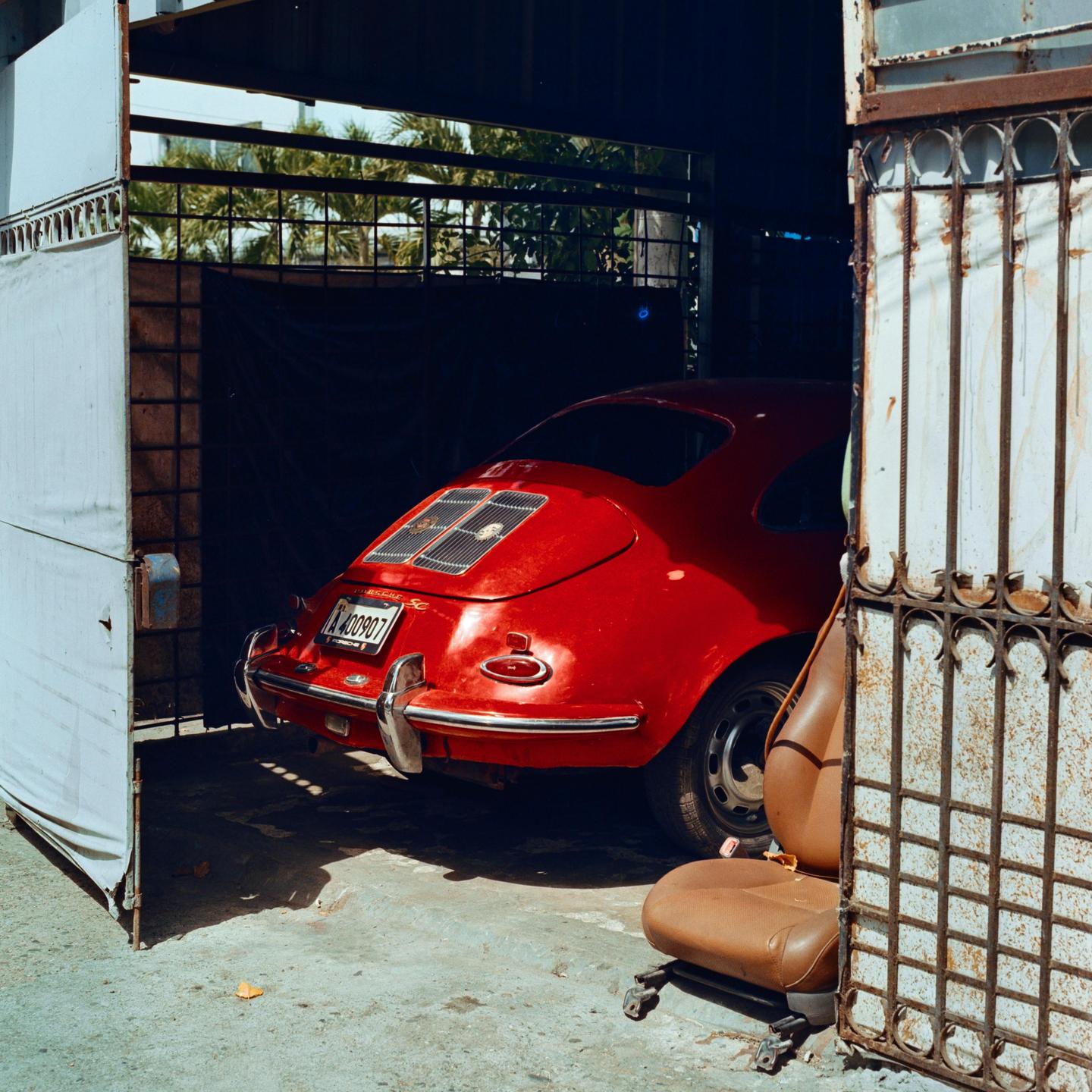 A red vintage car is parked inside a garage with its gate partially open.
