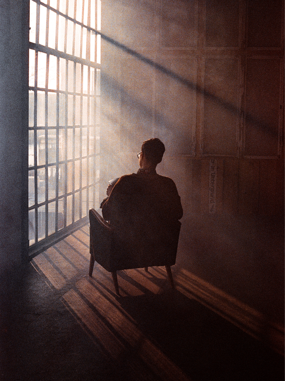 A man sits in an armchair, looking out a large window with bars, with light streaming through the window and casting shadows on the floor.