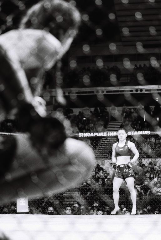 A woman stands in a fighting ring while another fighter is blurred in the foreground.
