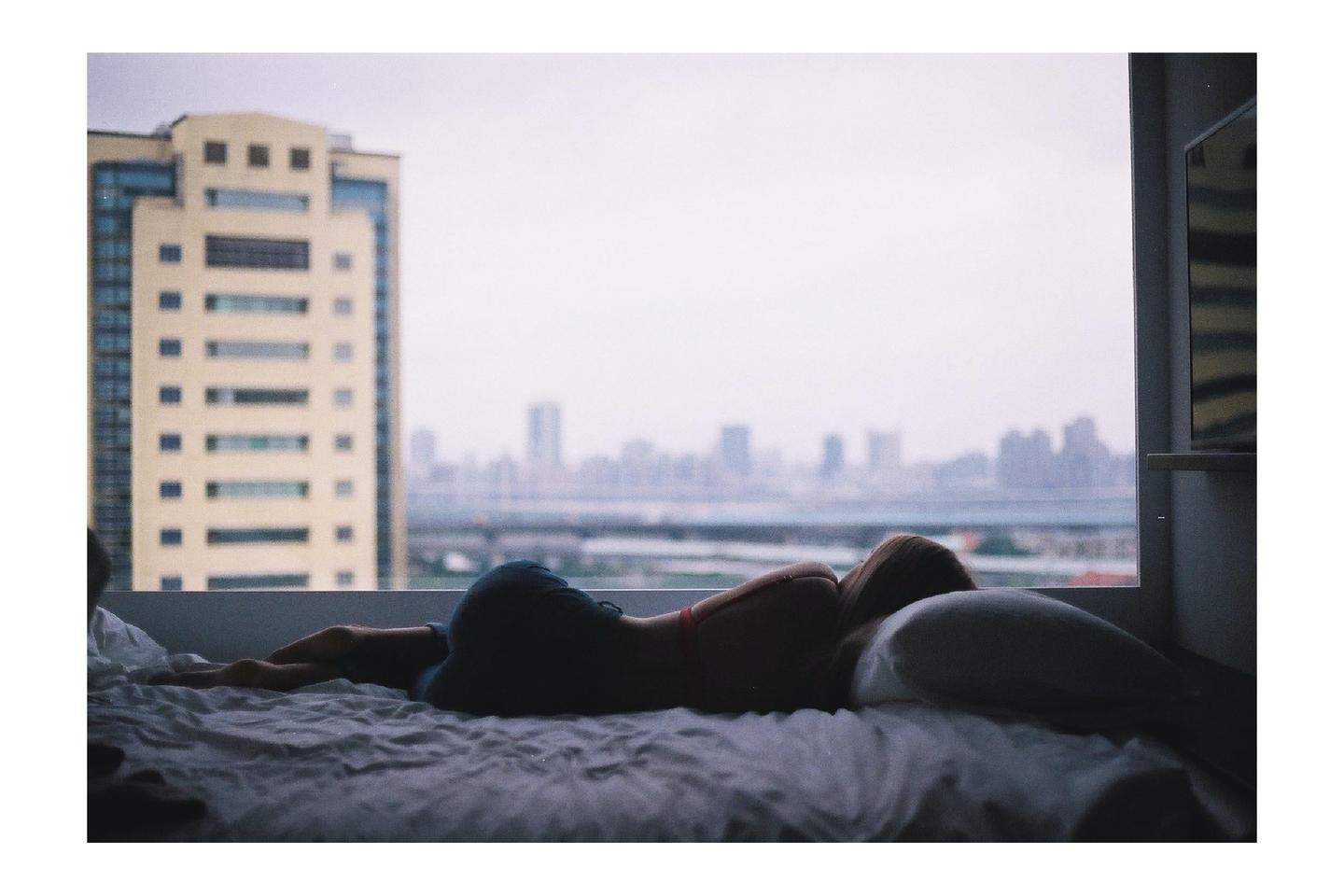 A woman lies on a bed looking out a window at a city skyline.