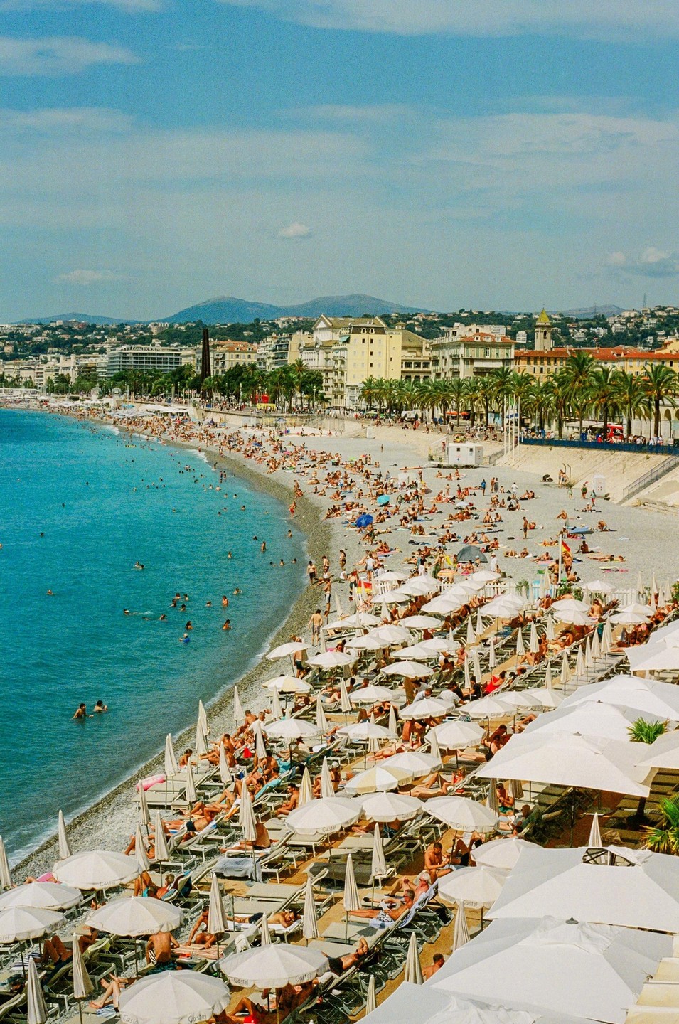 A crowded beach with many people swimming and sunbathing under white umbrellas is visible next to a city with buildings and palm trees.
