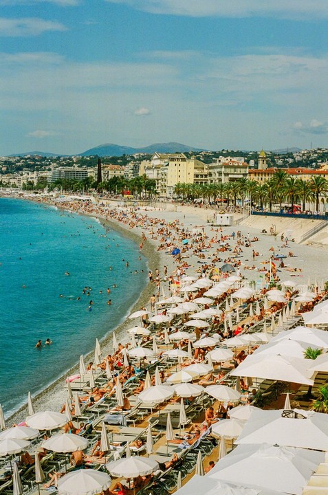 A crowded beach with many people swimming and sunbathing under white umbrellas is visible next to a city with buildings and palm trees.