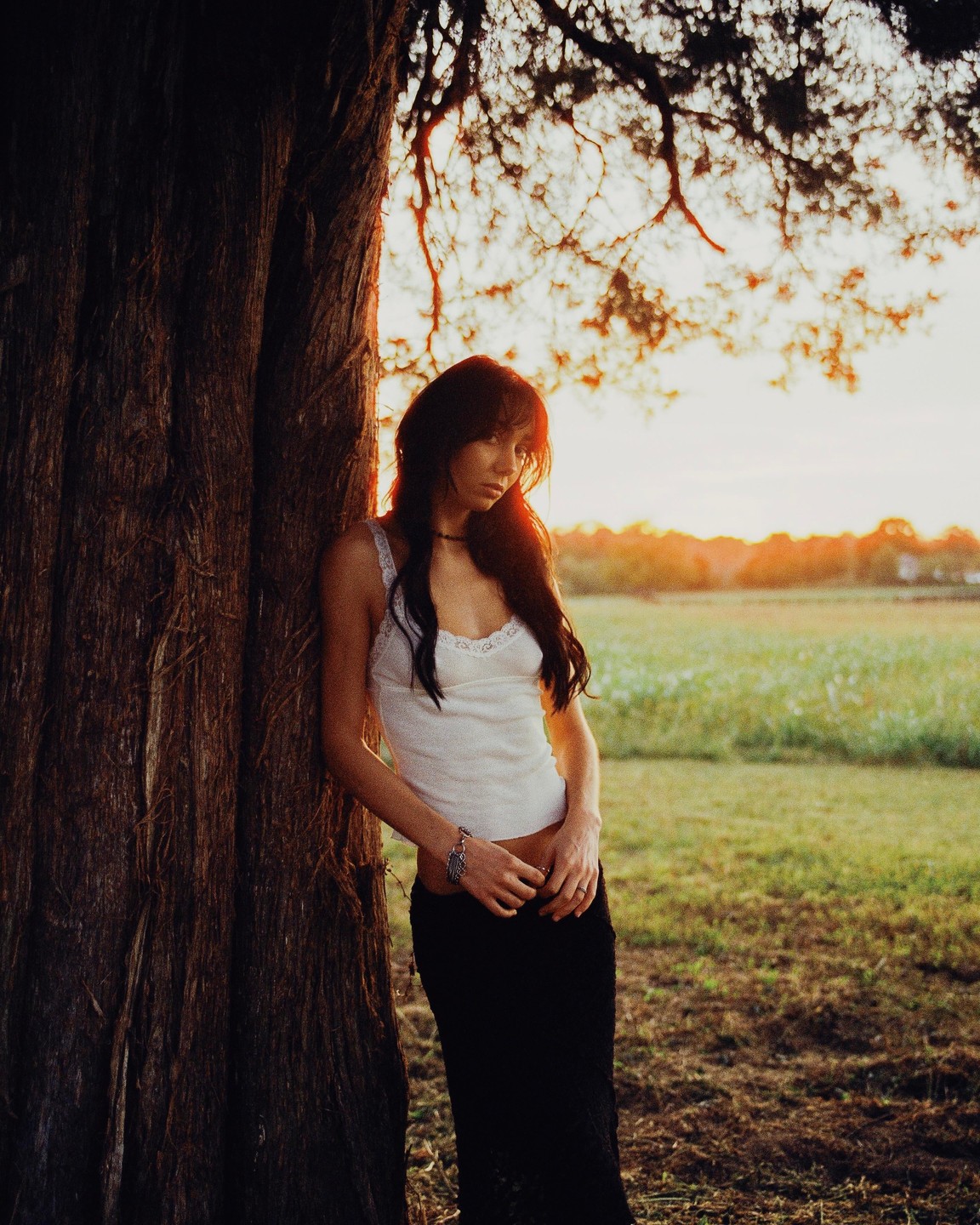 A woman with long dark hair leans against a tree trunk in a field at sunset.
